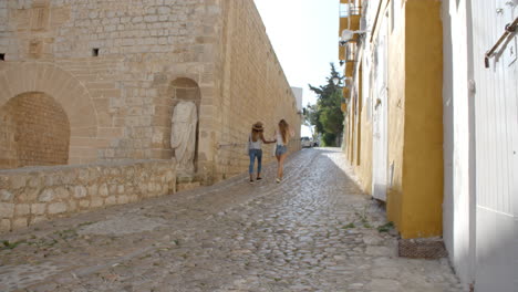 Two-female-friends-walking-through-old-Ibiza,-back-view